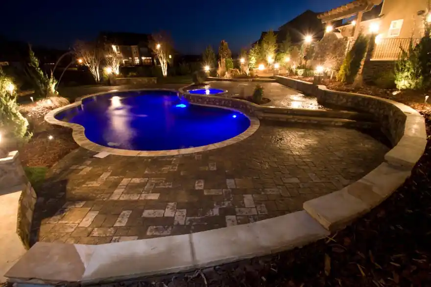 Paver deck and stone coping around a blue-lit pool at night with uplit trees