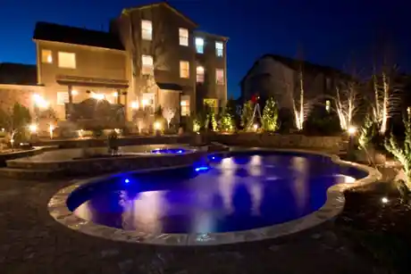 Freeform pool with stone coping and blue LED lights, a raised spa behind it and uplit shrubs along a paver deck at night