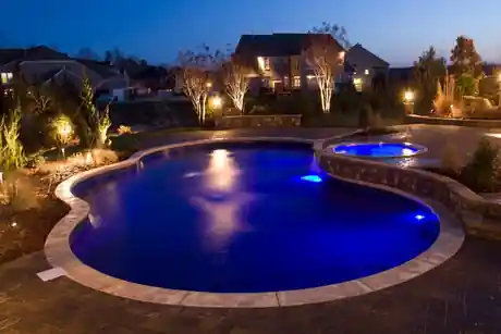 Kidney-shaped pool with pale coping, a lit round spa at the far end and uplit birches at dusk